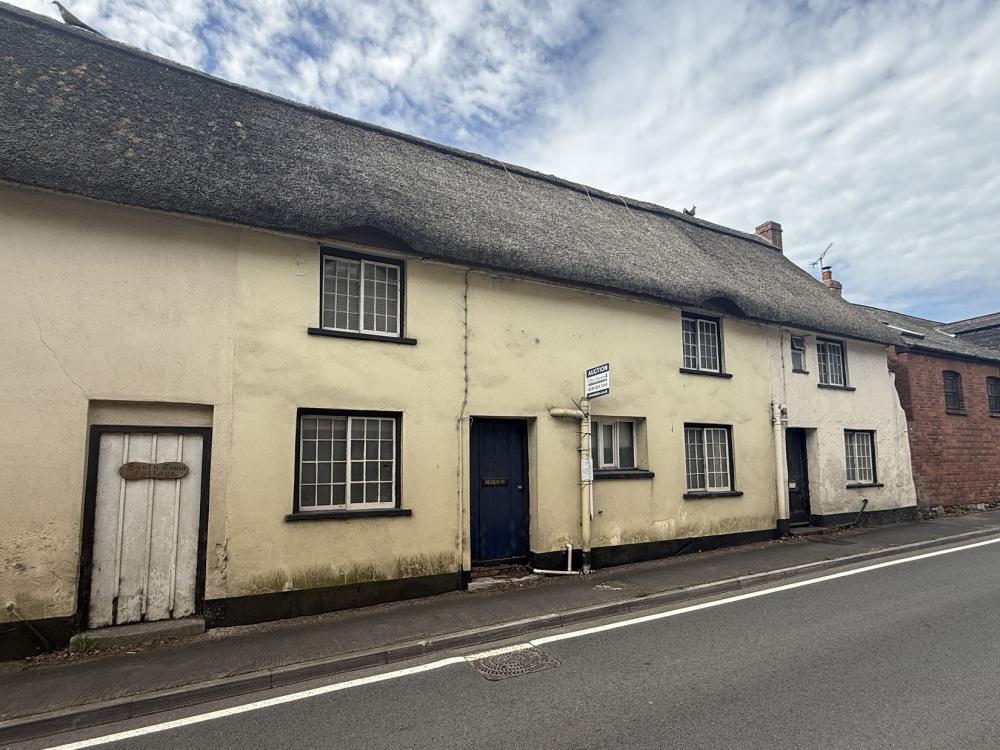 the image shows a mid terraced cottage with faded yellow exterior under a thatched roof. The property has a front door in the middle and is flagged with single glazed white framed windows on either side and above on the first floor. The property sits by the road. The sky is mostly white cloud with some flecks of blue.
