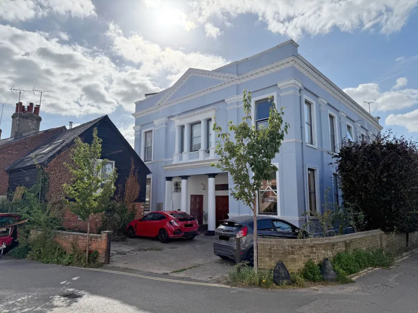 Large detached pale blue building with two white pillars at the entrance. Sitting on the corner of a road with off street parking in front of it and a red car and blue car parked. Sky is blue with white clouds and sun peaking through.