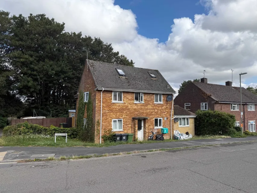 Detached brown brick house under pitched roof. Sitting kerbside with road in front and trees behind. The sky is white clouds with some blue poking through.