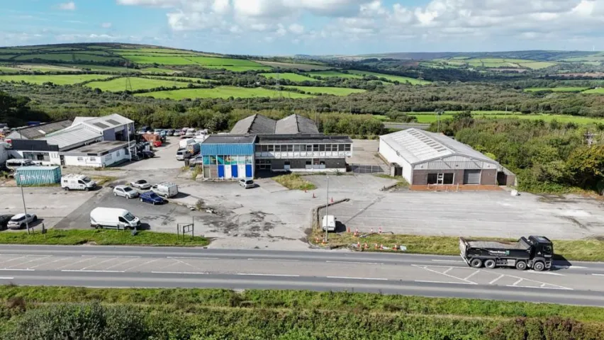 the image shows a large industrial park in the foreground with gree fields and hills and trees in the background. The buildings sit on a grey concrete forecourt. There are a handful of white cars and two white vans parked,