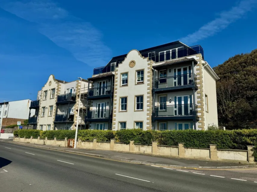 the image shows a substantial four floor building with several windows and double doors leading to a balcony. The property has a green hedge running along the front and there is a partial view of the grey road. The sky is blue.