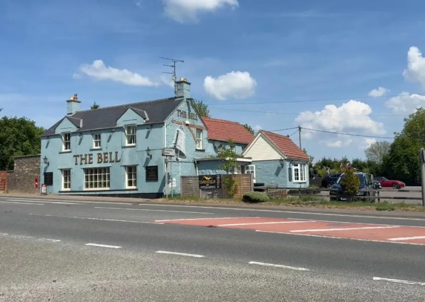 The image shows an pale blue detached building with The Bell displayed on the front external wall and the side. The property sits by the road and there is a car park to the side with a black car parked. The sky is mainly blue with a few white clouds.