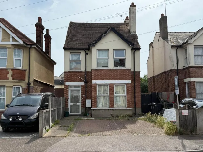 A detached house with brown brick cladding on ground floor, two white framed windows and a white framed glass front door. The first floor is painted light yellow and three white framed windows. There is a paved driveway in front of the house, The sky is mostly white with flecks of blue,