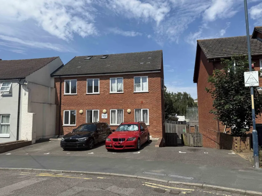 A brown brick built property with four sets of white framed windows on both the ground and first floors. There is off road parking in front of the property with a red car and black car parked. The driveway leads straight on to the road. The sky is blue.