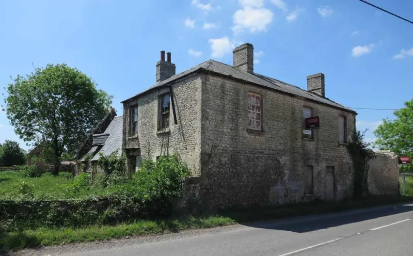 Detached brick house that looks derelict. Surrounded by overgrown greenery and sitting by a road.