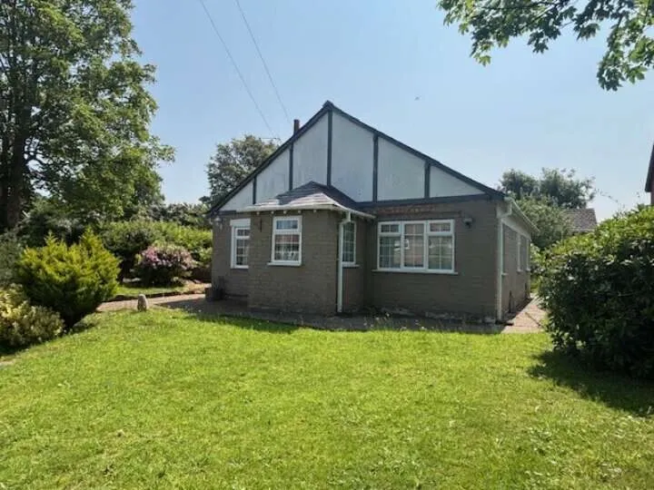 A detached bungalow with brown brick exterior and a white pitched roof sitting on a large green grass plot. The sky behind is blue.
