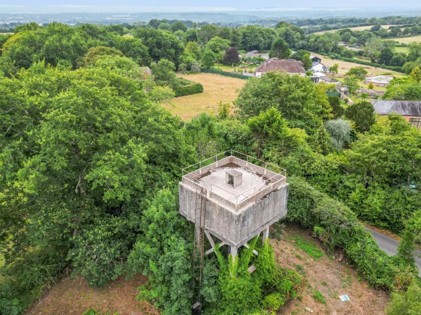 A disused former water tower in the centre of the image surrounded by green bushes and trees and a scattering of houses in the distance