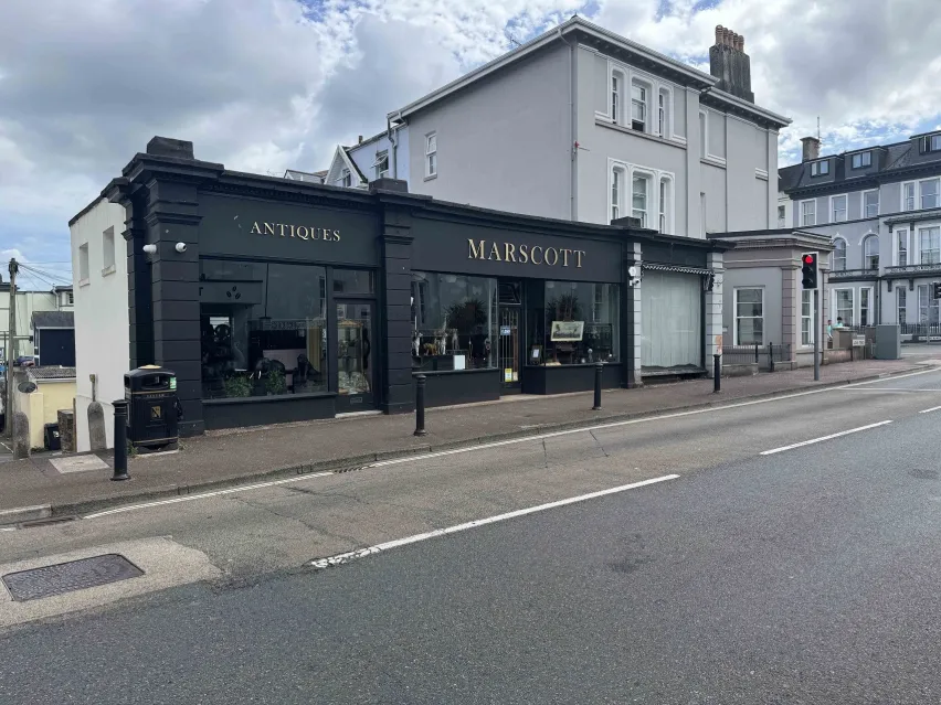 A single storey commercial property with black exterior and gold lettered writing. The property is attached to a three storey white building and both site kerbside next to a road.