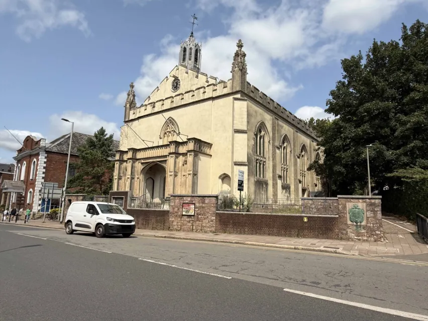 A large cream coloured detached building that resemble a church with large windows on the side and columns to the front. The sky is blue with white clouds. There is a road in front with a white car parked.