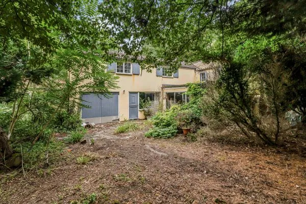 the image shows glimpses of a detached house with grey colour garage. There are lots of green trees in the foreground and a brown soil grassland in front of that.
