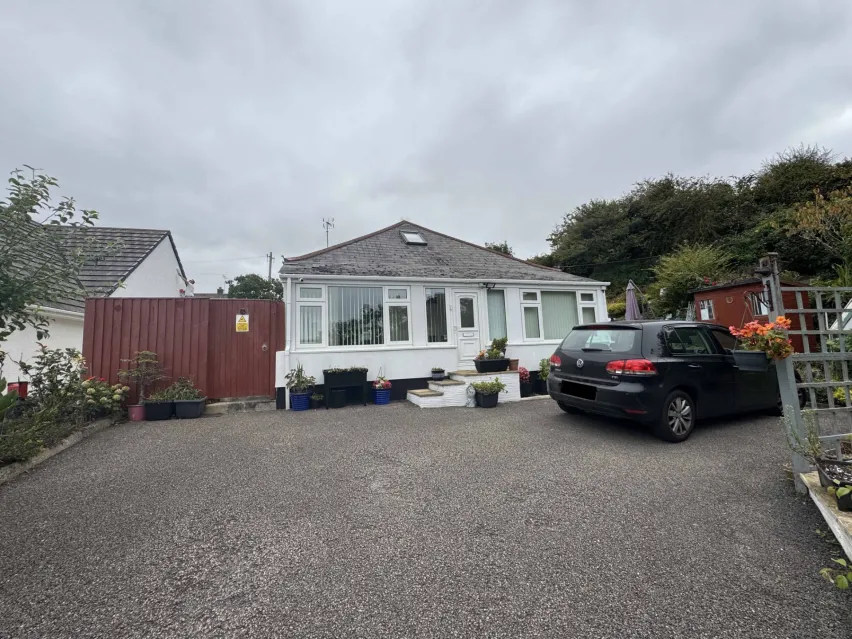 a white single storey bungalow with large front facing windows sitting on a tarmac driveway with one black car parked up