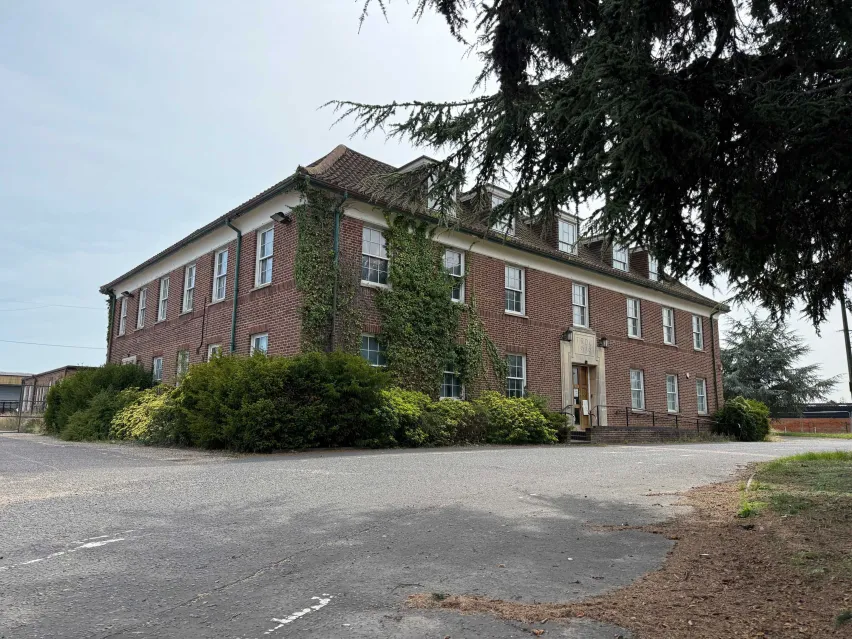 A substantial brown brick detached building sitting alongside a grey tarmac road and surrounded by low level green bushes