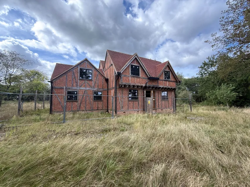 A detached brown brick house with side extension stting in an overgrown grass plot. The sky behind is blue with large grey and white clouds.