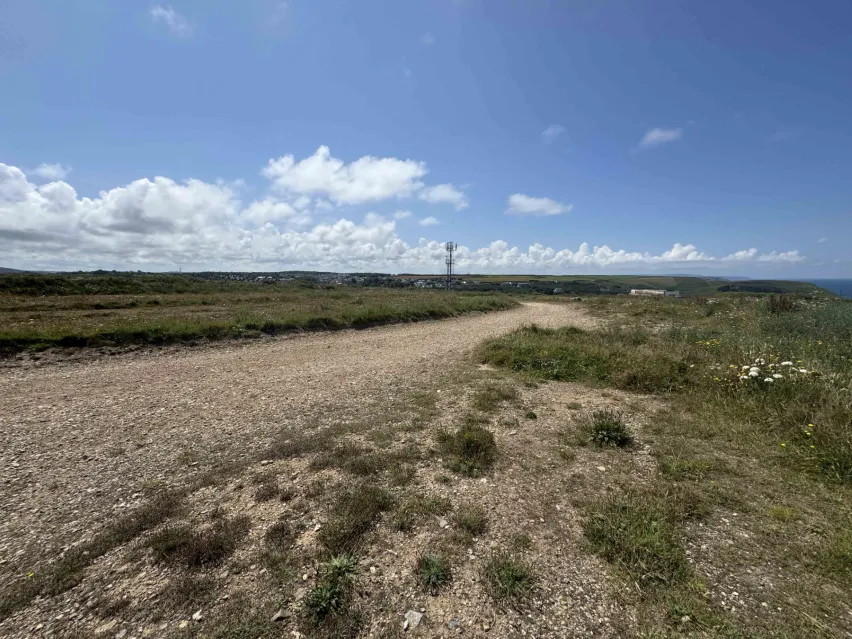 a strip of gravel land surrounded by green grass and fields with partial glimpse of the sea. The sky is blue with a few white clouds.