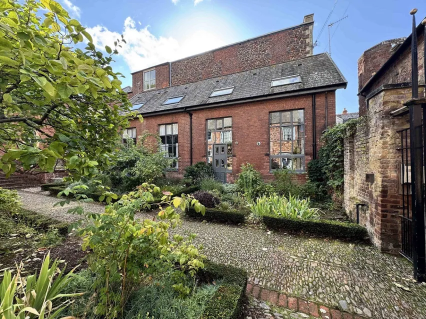 Brown brick building with large windows sitting behind a gravel pathway with green plants and a tree in the foreground