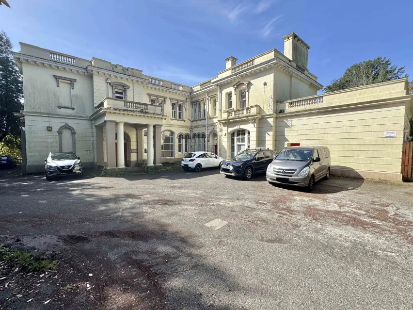 A large detached cream coloured mansion house with pillars at the front door.  The building sits on a large car parking area with four cars parked. The sky is blue.