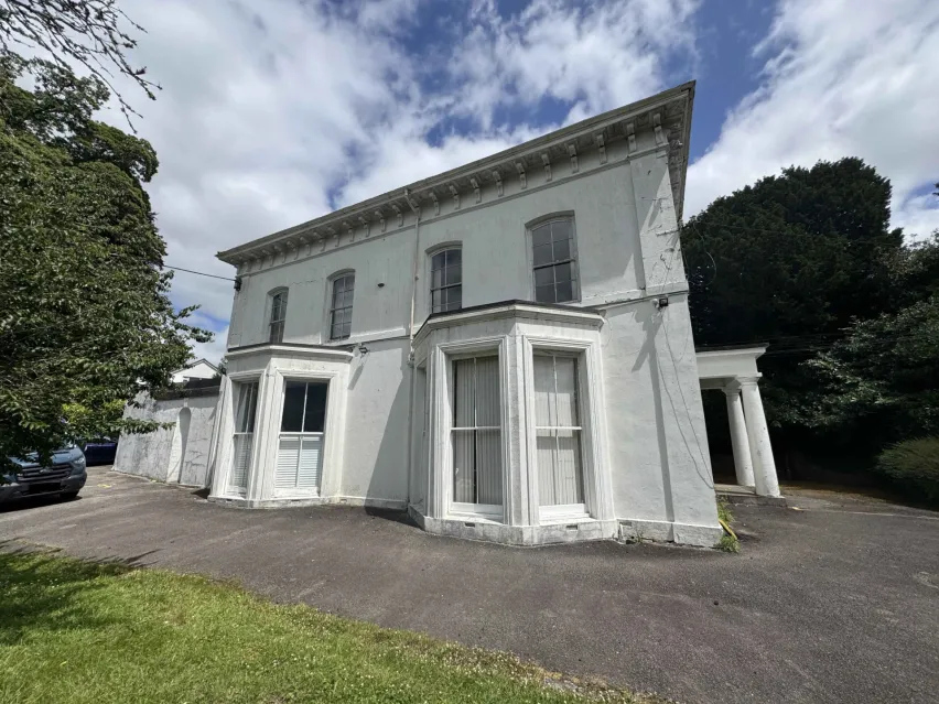 Detached white residential villa with two large bay windows on the ground floor. Sitting on tarmac with glimspes of green grass in front and trees behind, with white cloudy blue sky in background.