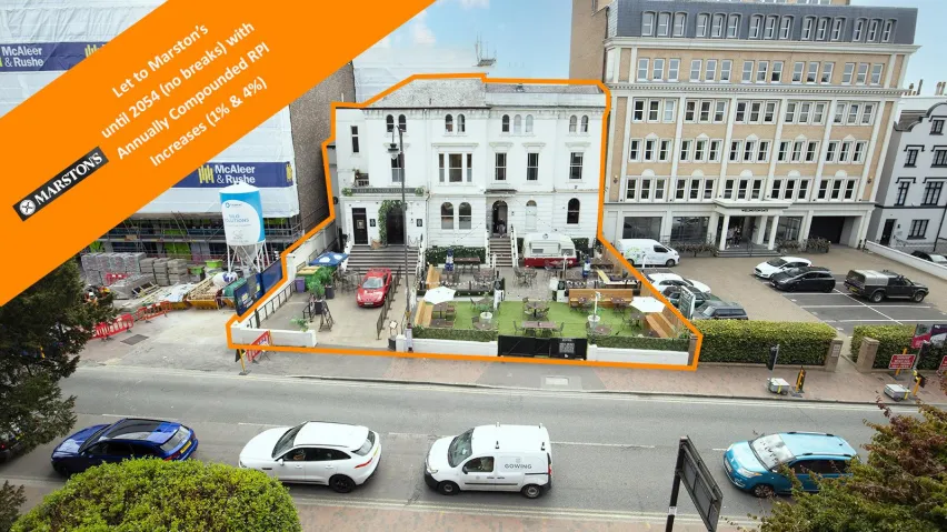 Large white detached public house buiilding set back behind a main road and surrounded by buildings. 