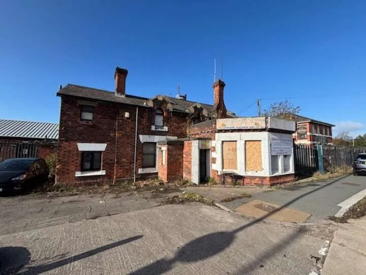 A substantial detached brown brick building with boarded up windows. Blue sky behind and sitting on a tarmac parking area.