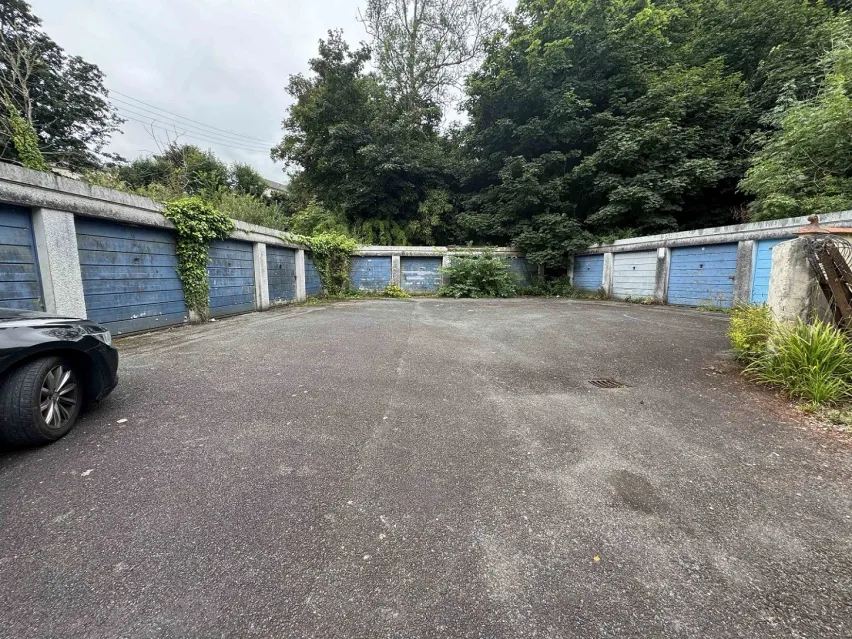 large grey tarmac forecourt with a rown of white and blue door single lock up garages on either side. There are green trees behind the garages and a patch of blue white sky.