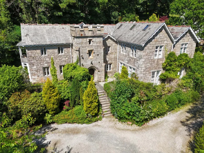 A large detached grey stone building on two floors surrounded by green bushes and trees.