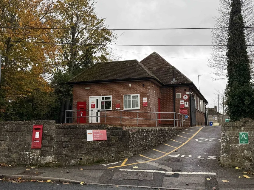 Detached brown brick building alongside a tarmac road, situated on a corner plot. The sky is grey and there are a couple of trees next to the building with orange and green leaves.