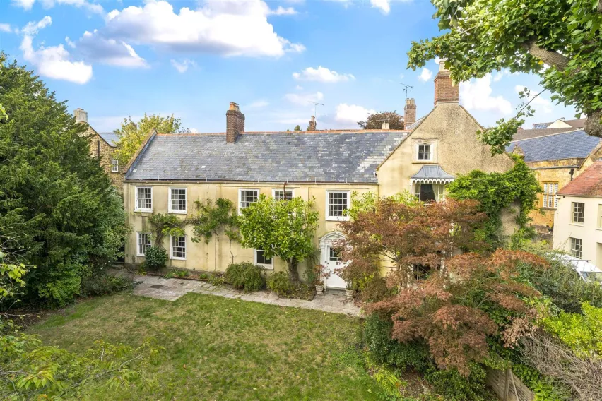 Partially obcured by greenery and trees, a yellow sandstone house on two floors sitting back behind a green grass garden with trees and bushes. The sky is blue with white clouds.