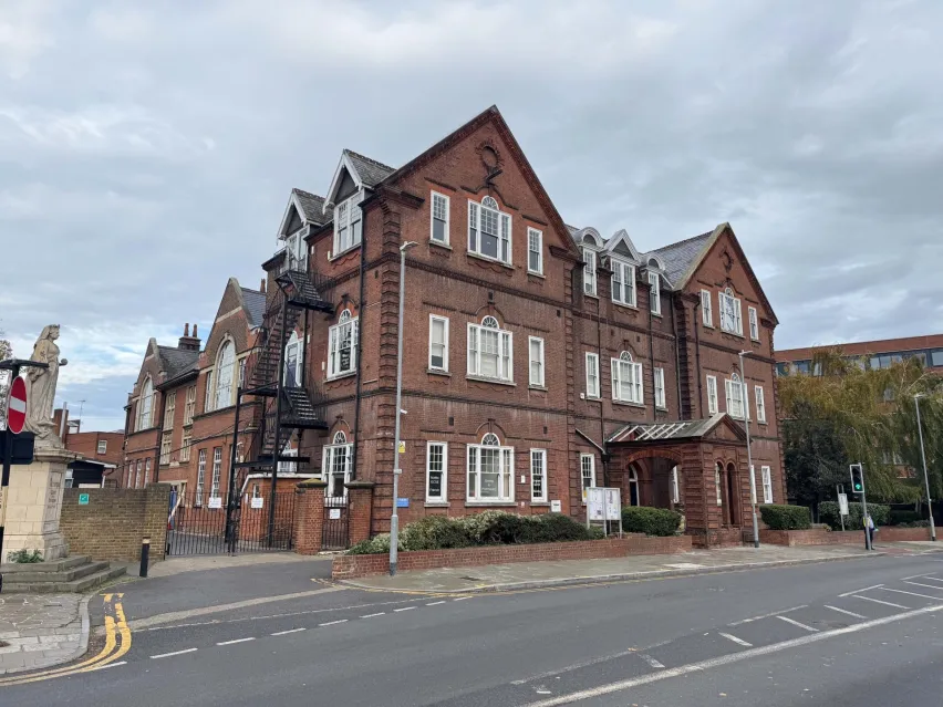 Substantial detached three storey brown brick building with multiple white frames windows on each floor. Alongside grey tarmac road. The sky is mostly grey and white cloud.