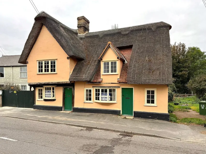 a pale orange painted detached thatched property with two green front doors set next to a road.