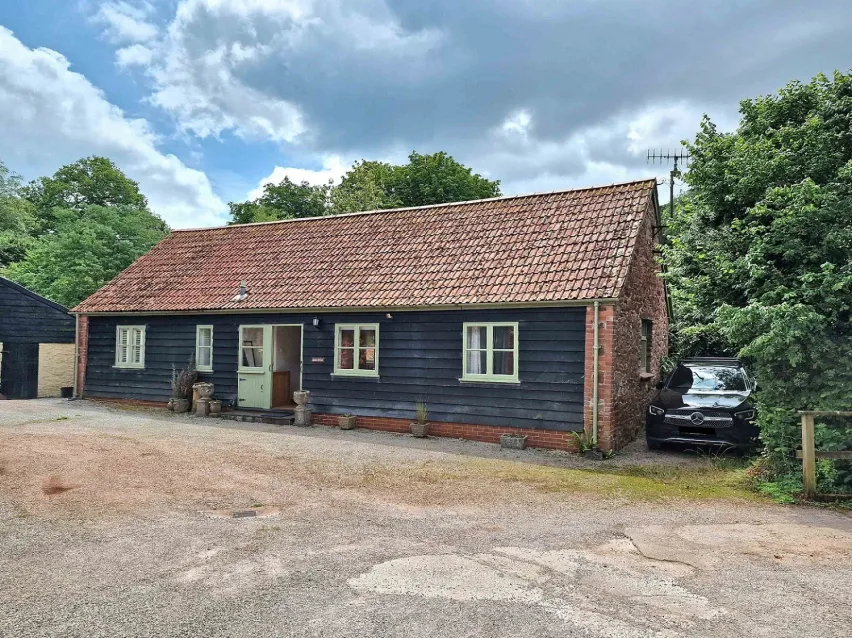 Detached single storey residential property  surrounded by green trees and sitting on a paved forecourt. The sky is blue with grey and white clouds.