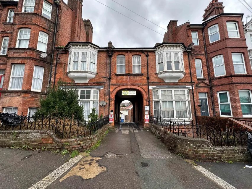 Brown brick buildings with multiple white framed windows. A mix of two and three storey properties sitting alongside a grey tarmac driveway. The sky is grey and cloudy.
