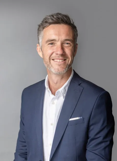 Middle aged white gentleman smiling at the camera. He is wearing a navy blue suit jacket and white open necked shirt.