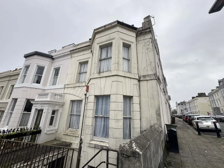 A dirty, cream coloured, end of terrace residential building. There is a pavement along the right hand side and the sky above is white cloud.