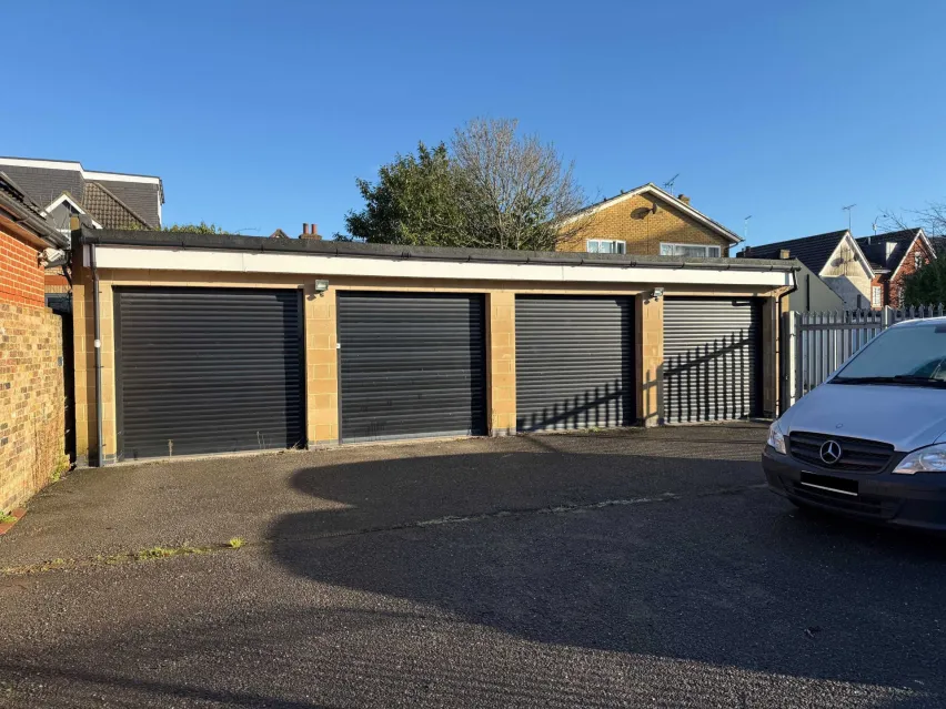 a row of four single garages with black doors, on a grey tarmac hardstanding. Blue sky with tops of houses in the background.