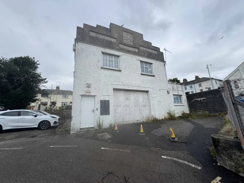 A detached white building with grey roof and grey framed windows sitting in a tarmac car park. The sky is mainly white and grey and there is a white car parked on the left.