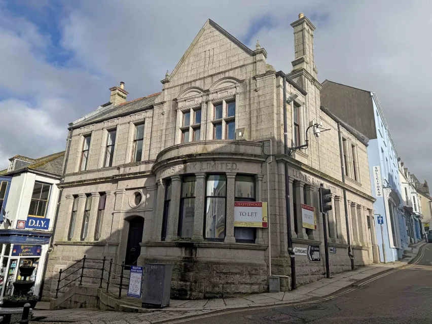 A light brown brick building on the corner of a junction. Mulitple windows on both ground and first floors. The sky is white cloud with glimpses of blue. 