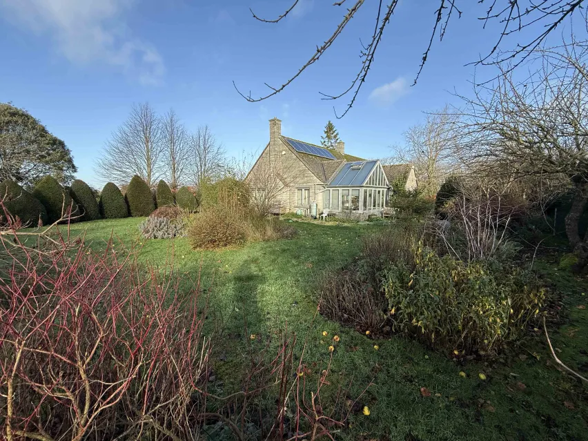A stone built bungalow set in a large garden with green grass and bare leaf trees, The sky is blue with a couple of white clouds.