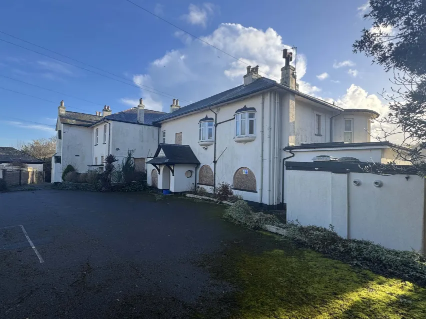 A large white detached property with white garages next door. Sitting on a tarmac driveway with blue sky above.