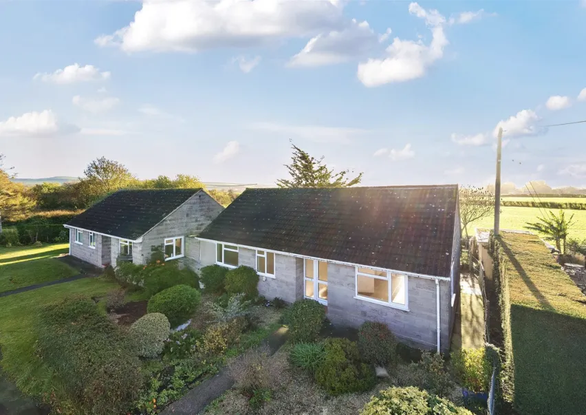 Two grey brick bungalows side by side sitting on a green grass plot with green fields behind, blue sky with white clouds.