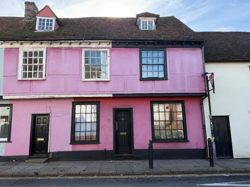 A mid terrace bright pink period building that appears to have a ground floor, first floor and a room in the upper parts. It sits kerbside and the sky is blue with streaks of white cloud.