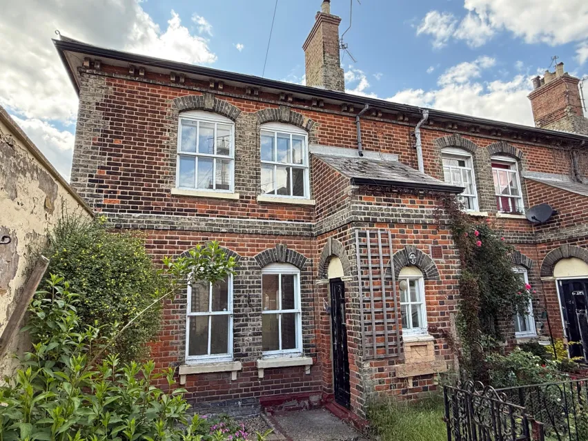 An end of terrace brown brick house with a pair of white framed windows on both the ground and first floor. There is a green bush in front and the sky is blue with white cloud.