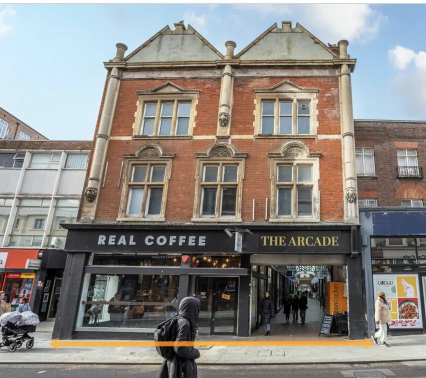 A mid terraced brown brick, three storey building next to road. There is a coffee shop on the ground floor and entrance to The Arcade shopping centre. On the first and second floors there are three sets and two sets of windows respectively. 
