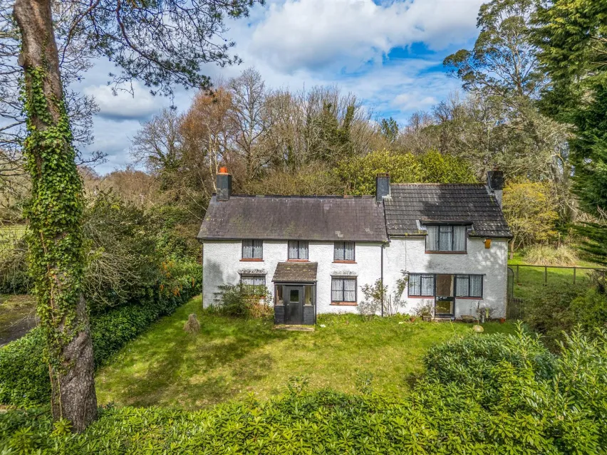Large detached white house under pitched roof sitting in glorious green gardens with blue sky dotted with white clouds