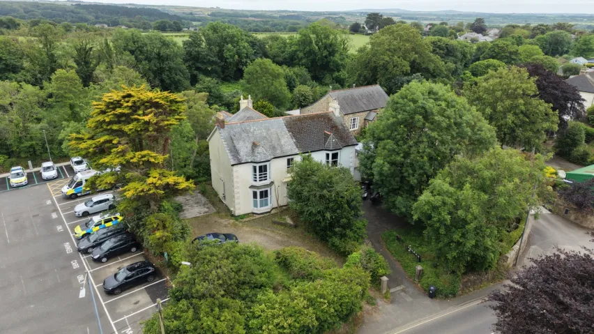 An aerial view shot of a white semi-detached house surrounded by green treess and bushes with a tarmac car park to the left side