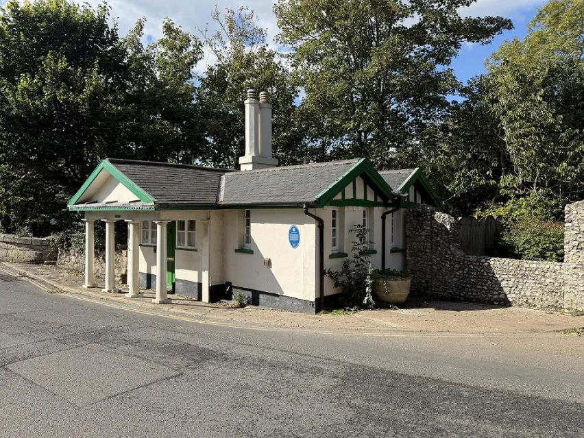 A detached single storey toll house with pillars on the outside. Brick wall and green trees in the background with glimpes of blue sky.