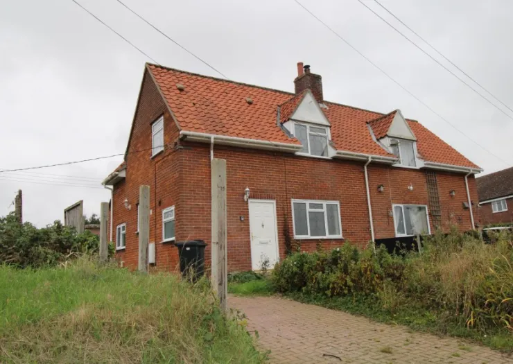 Red brick house with tiled roof, white door, and paved driveway. Overgrown garden.