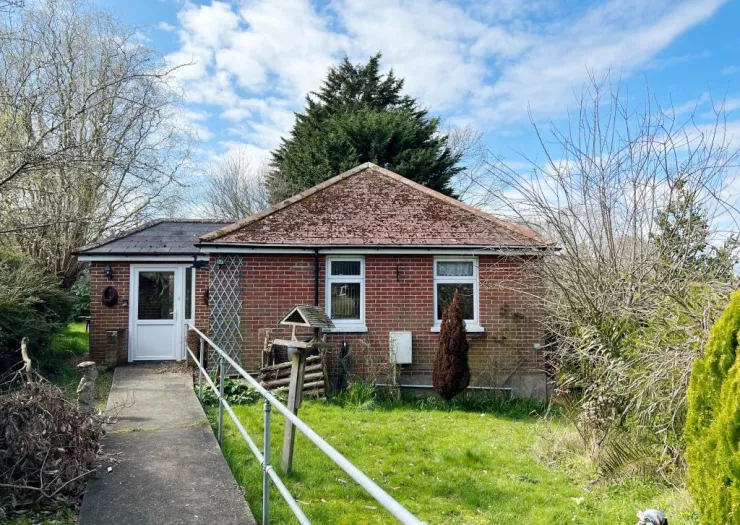 Red brick bungalow with a sloped tiled roof, white-framed windows, accessible ramp, and a grassy front yard.