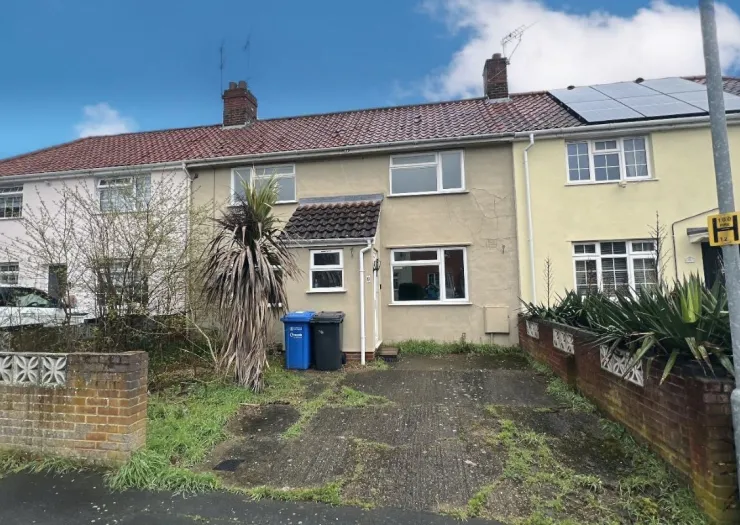 Tan terraced house exterior with red tiled roof, driveway, and overgrown front garden.