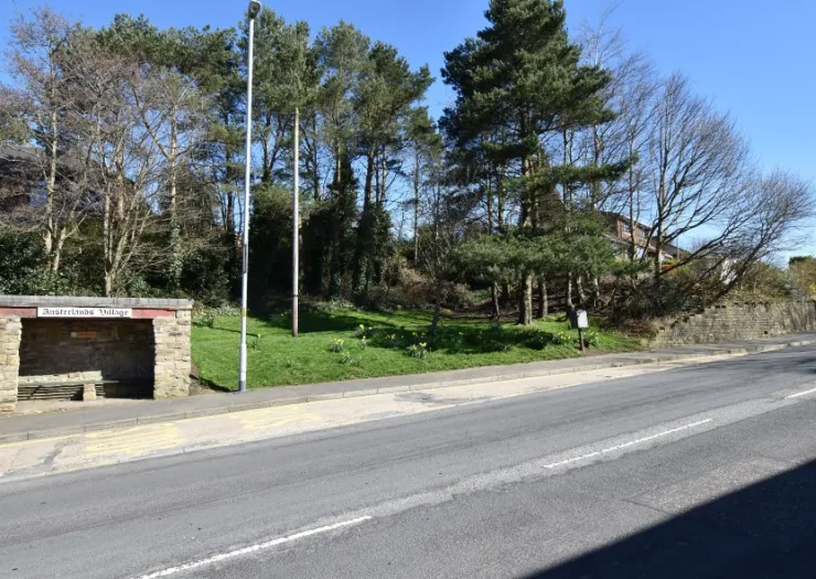 Austerlands Village bus stop with grassy area, trees, and stone wall on a sunny day.