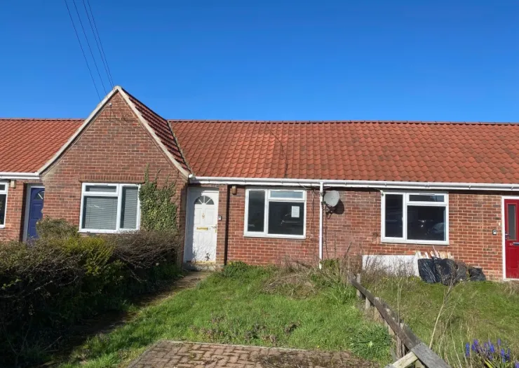 Brick bungalow with red tiled roof, white-framed windows, and overgrown front garden.